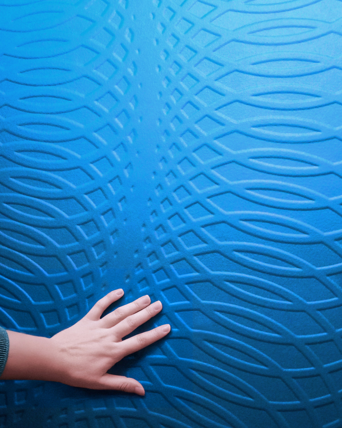 A close-up of a textured blue surface with an embossed wavy geometric pattern, with a hand resting on the lower left to show texture and scale.